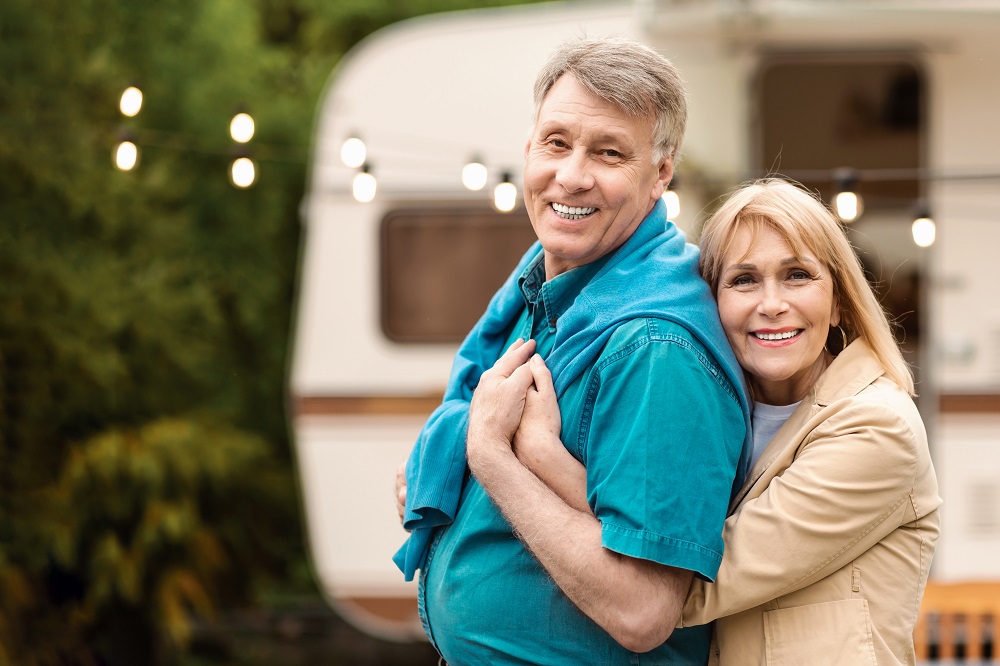 Camping together. Happy mature couple hugging each other on family vacation near their trailer. Panorama
