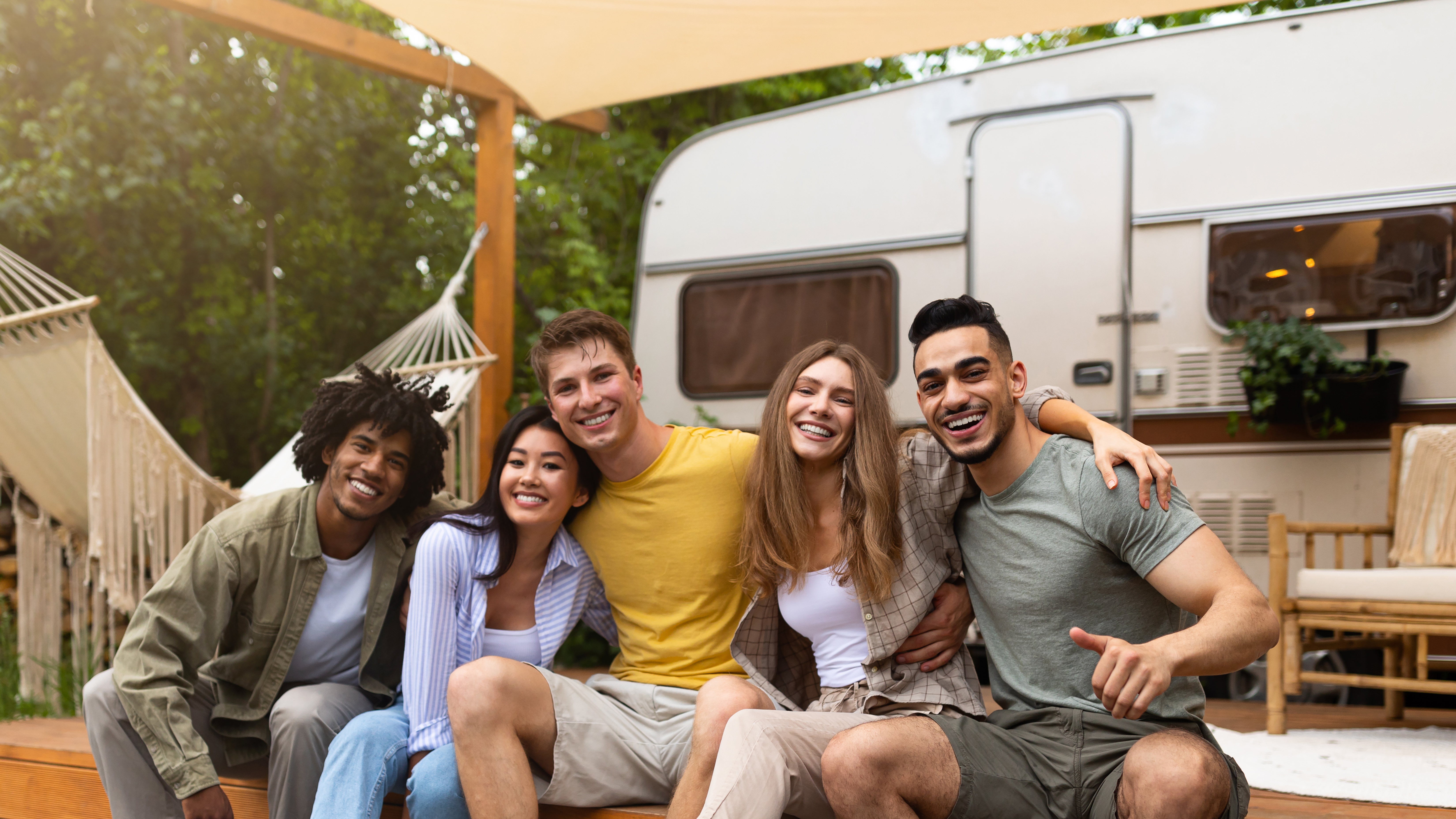 Full length portrait of multiracial young friends sitting near camper van, smiling at camera outdoors. Diverse millennials going on camping trip together, traveling in counryside